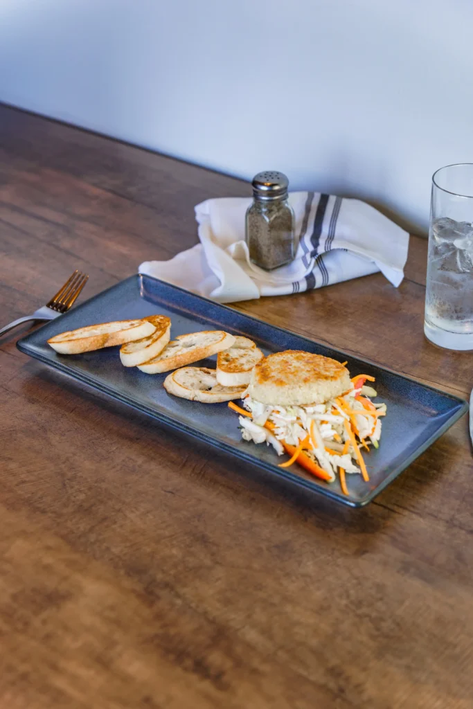 A rustic wooden table holds a rectangular plate with toasted baguette slices, coleslaw, and a patty. Utensil and drink nearby.